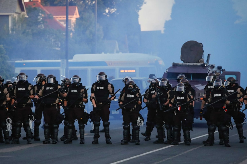 A row of heavily armed police stand in front of an armoured vehicle covered in smoke.