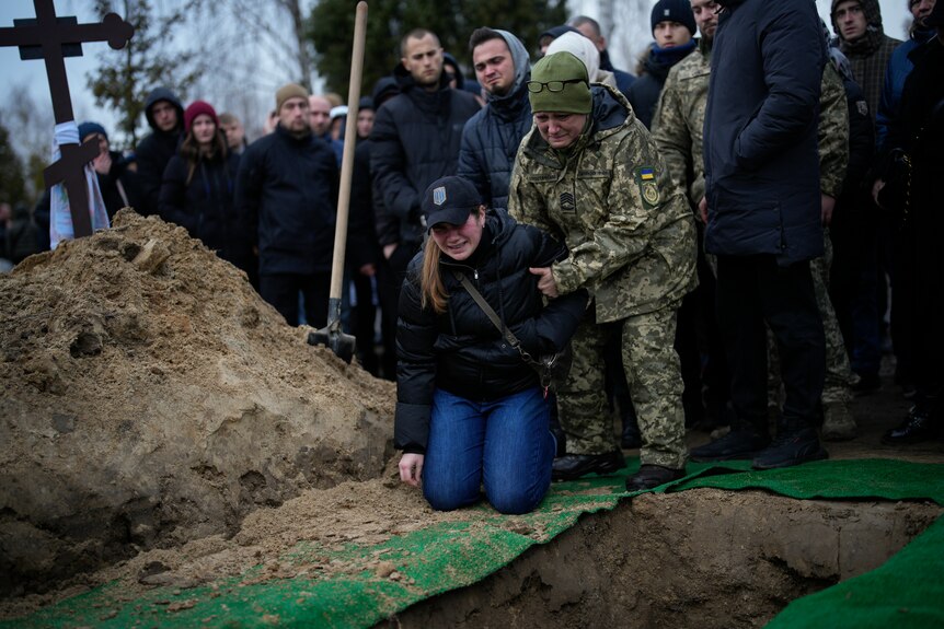 A woman is held by a Ukrainian soldier as she drops to her knees beside the grave of her fiance