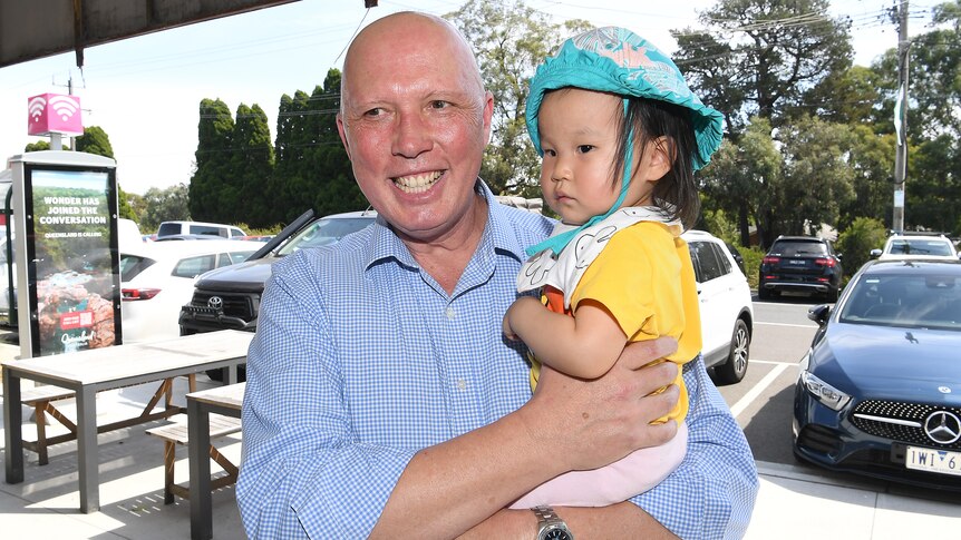 Peter Dutton holds a baby in his arms on a suburban sidewalk. 