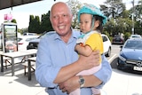 Peter Dutton holds a baby in his arms on a suburban sidewalk. 