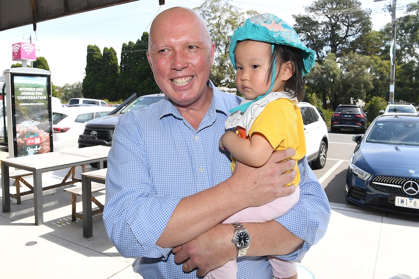 Peter Dutton holds a baby in his arms on a suburban sidewalk. 