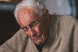 An elderly man in a tanned cardigan sits in a chair with his eyes closed against a dark background.