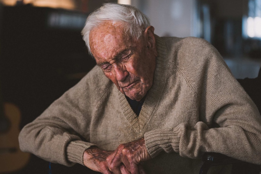 An elderly man in a tanned cardigan sits in a chair with his eyes closed against a dark background.