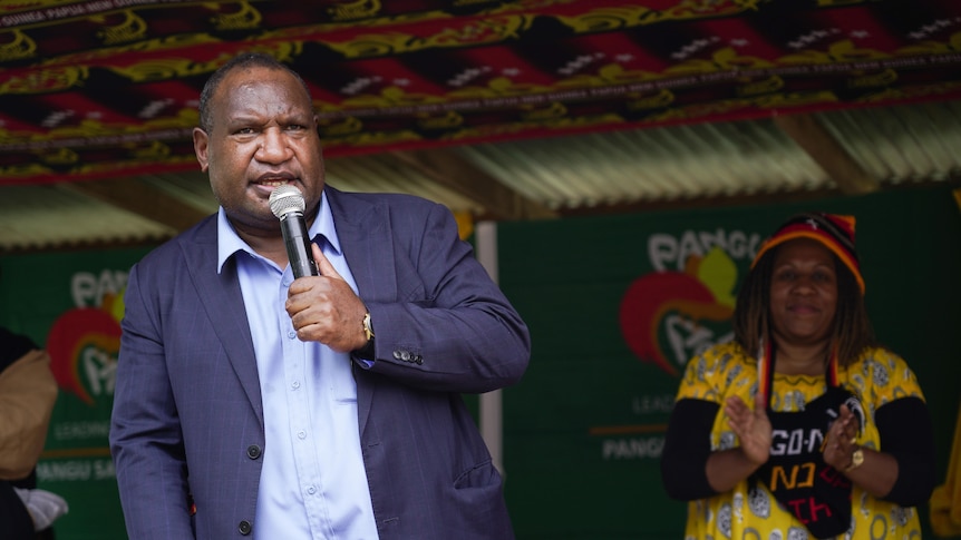 A man speaks into a microphone in front of a clapping supporter