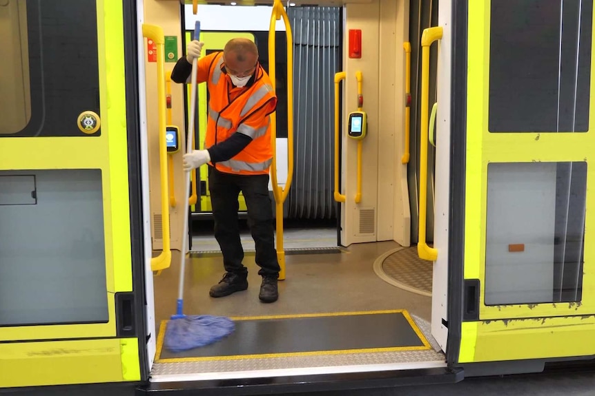 A man wearing a fluro vest, face mask and gloves mops the door of a tram.