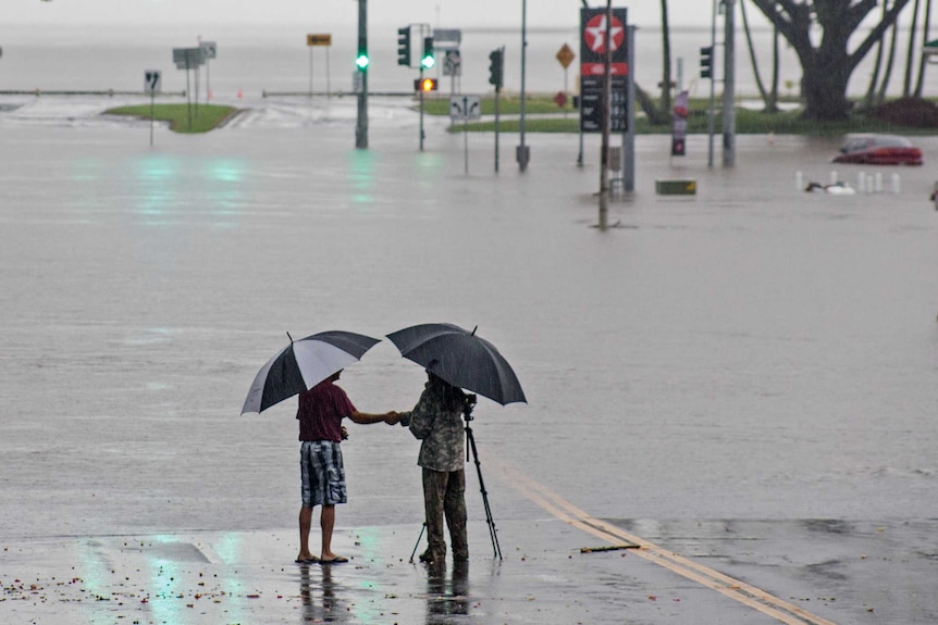 Two people stand under umbrellas at the edge of flood waters
