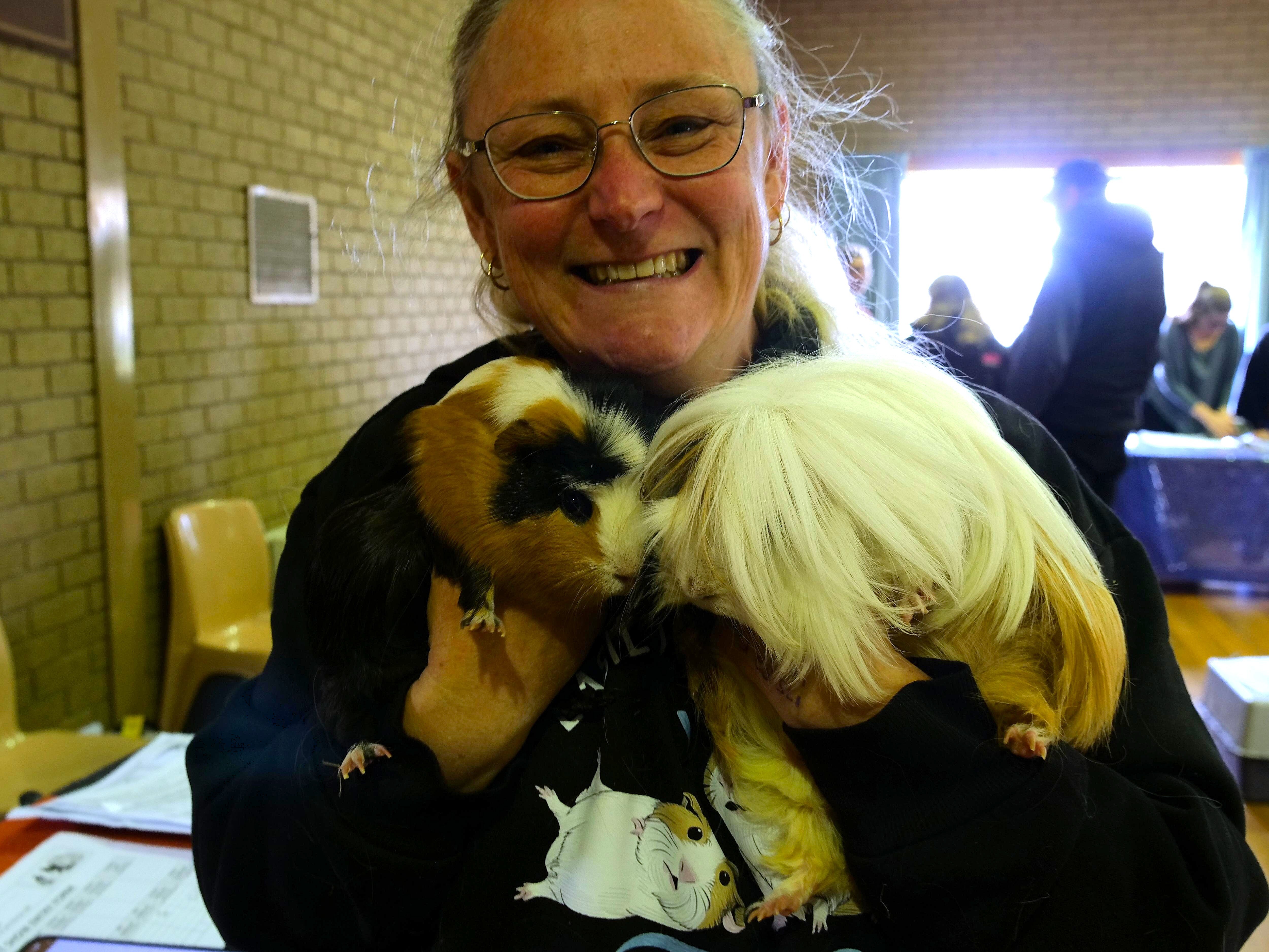 Uma mulher mais velha segurando duas cobaias contra o peito, sorrindo. 