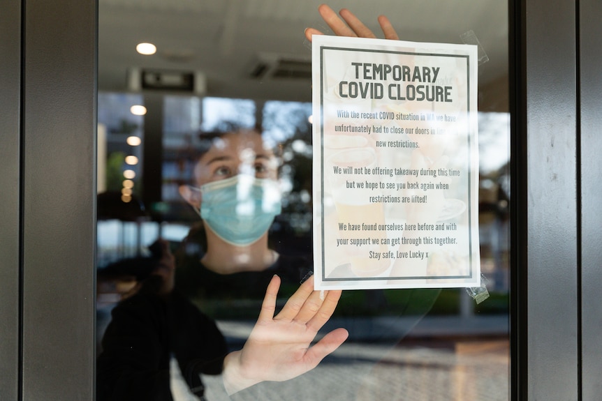 Woman in mask stands behind glass affixing poster