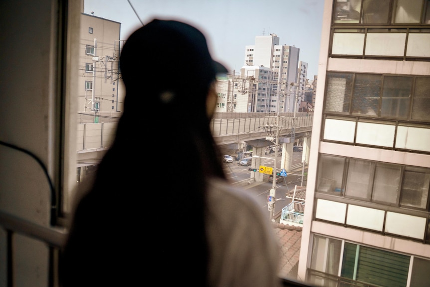 A woman with dark hair in a baseball cap looks out a window at a Seoul street