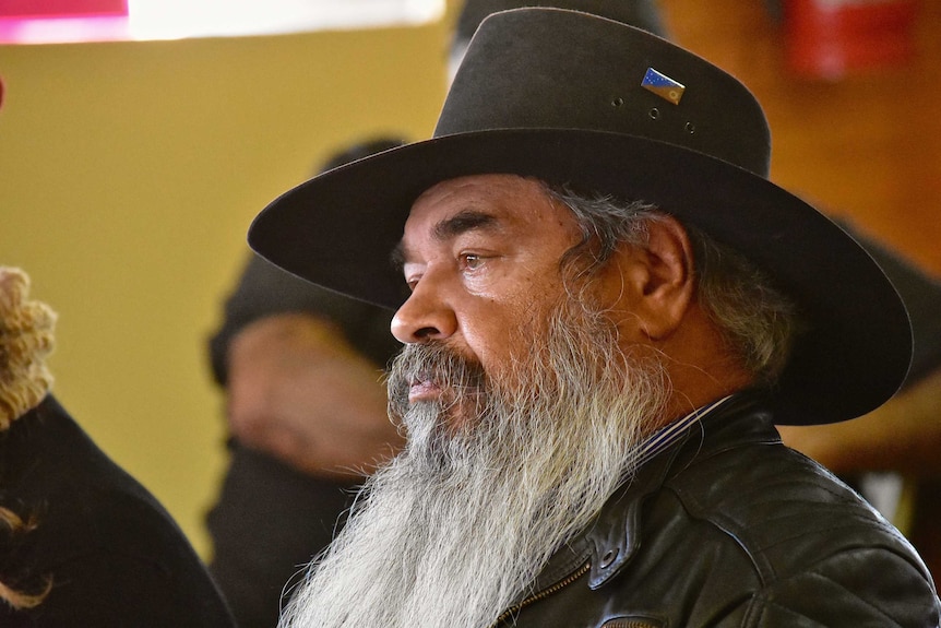 An Aboriginal man with a big white beard stares off camera.  He is wearing a wide brim hat