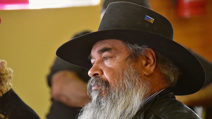 An Aboriginal man with a big white beard stares off camera.  He is wearing a wide brim hat
