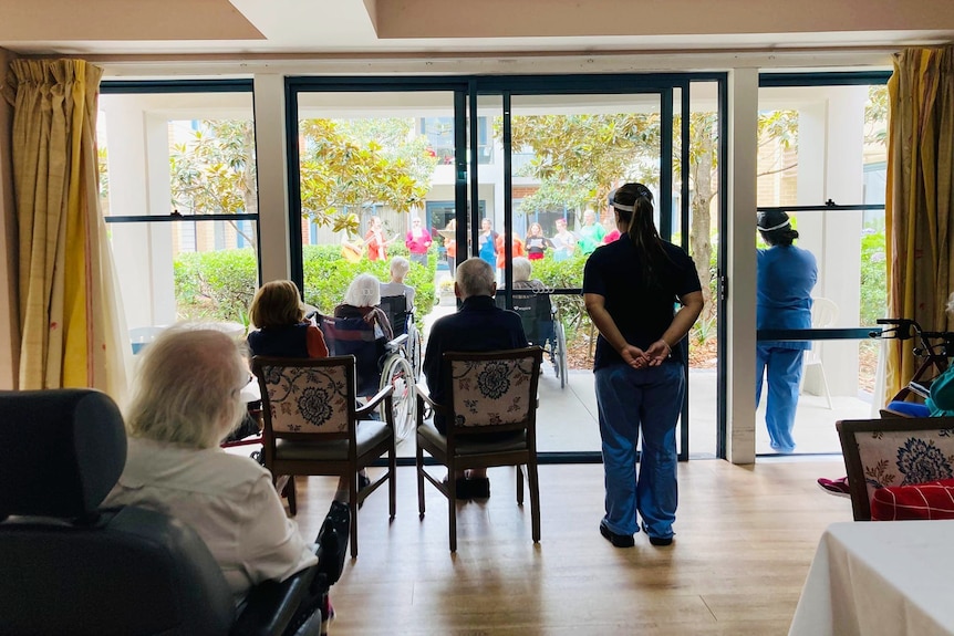 elderly residents at an aged care home watch people singing outdoors