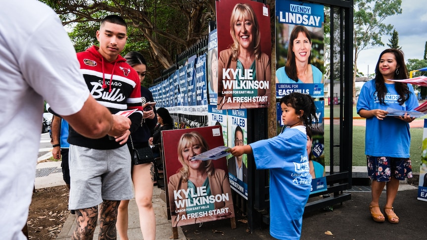a young child handing out how to votes at a polling booths with other poeple doing the same