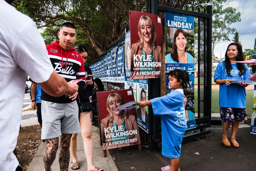 a young child handing out how to votes at a polling booths with other poeple doing the same