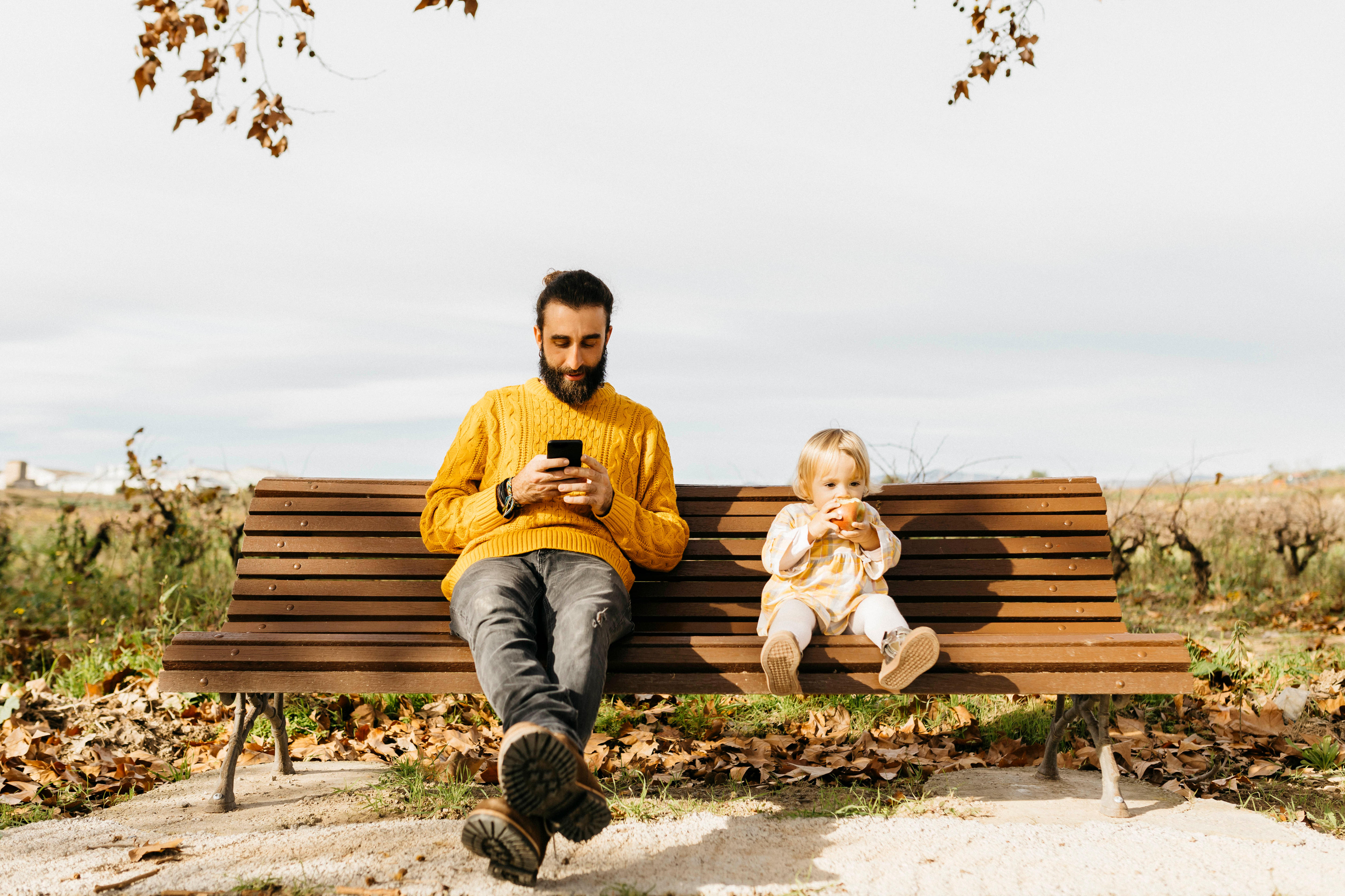 On a park bench, a seated man looks at his phone screen while a child sitting next to him stares ahead eating an apple.