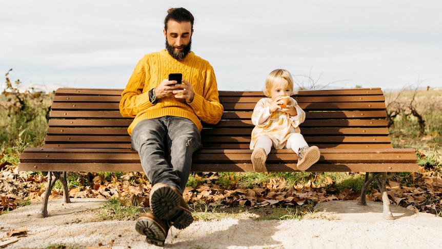 On a park bench, a seated man looks at his phone screen while a child sitting next to him stares ahead eating an apple.