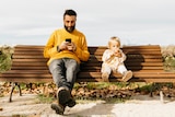 On a park bench, a seated man looks at his phone screen while a child sitting next to him stares ahead eating an apple.