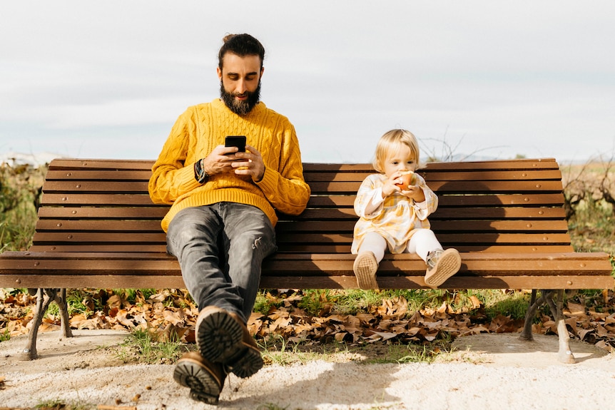 On a park bench, a seated man looks at his phone screen while a child sitting next to him stares ahead eating an apple.