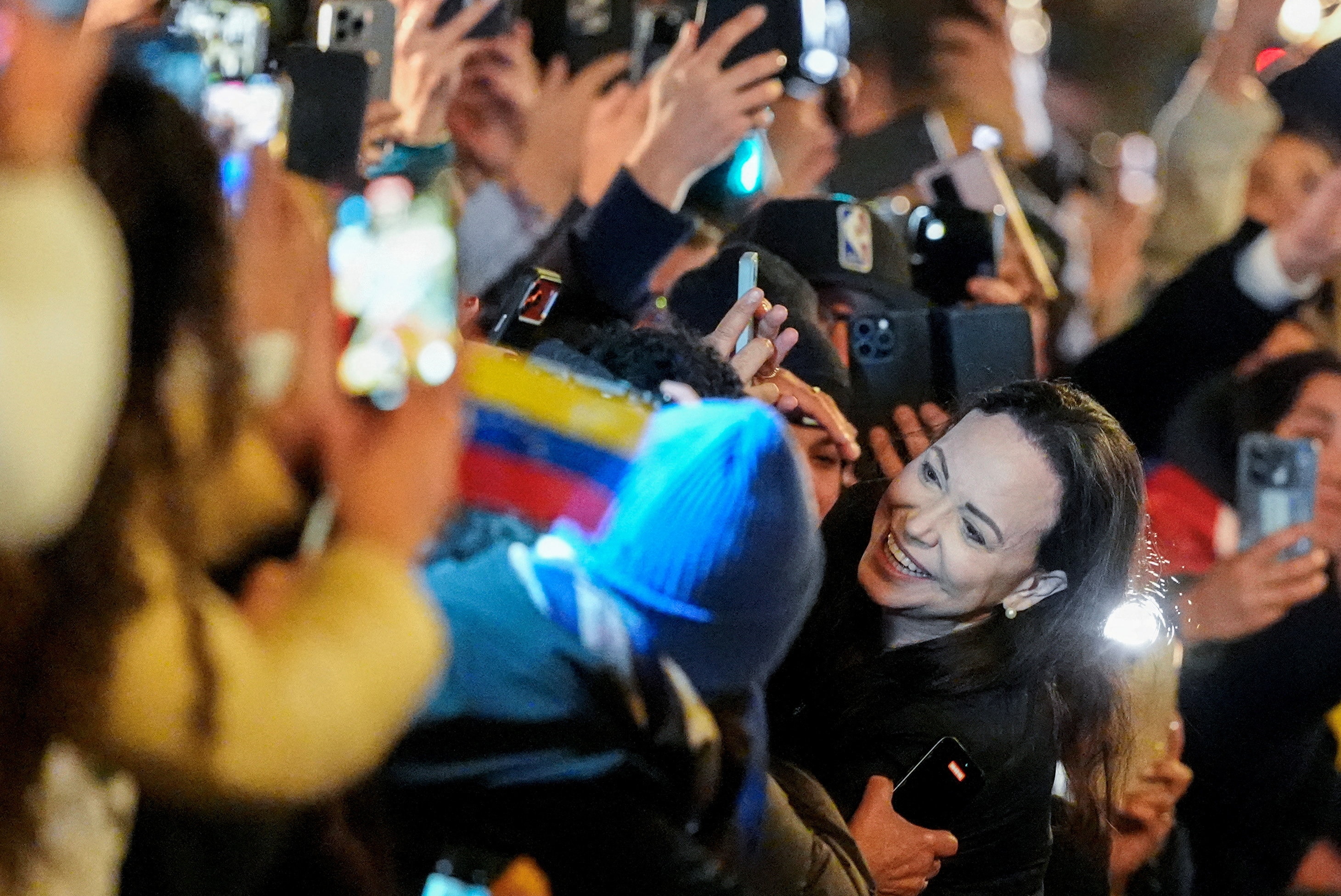 María Corina Machado sonríe y abraza a una gran multitud que ondea una bandera venezolana y sostiene teléfonos en el aire.