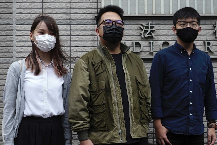 Bir kadın ve iki adam, gri tuğlalı adalet binasının önünde yüz maskeleriyle duruyor.