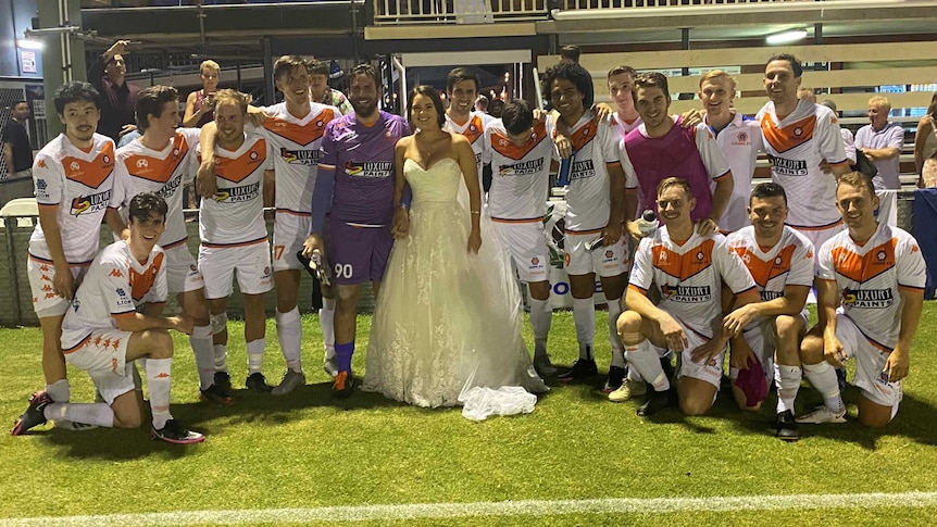 A woman in a wedding dress stands in the middle of the group of men wearing football kit in front of the clubhouse