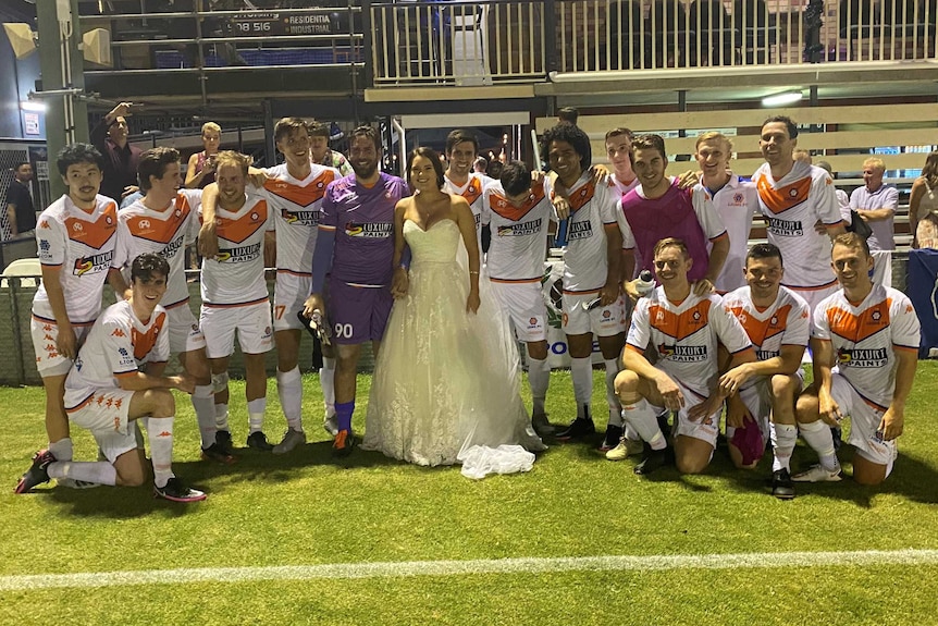 A woman in a wedding dress stands in the middle of the group of men wearing football kit in front of the clubhouse