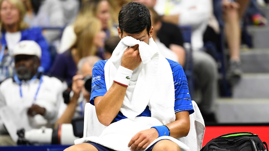 A male tennis player sits in a chair while holding his face in a towel.