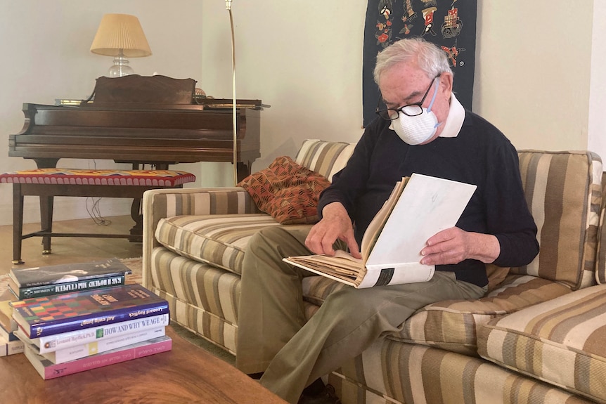 An elderly man wearing a surgical mask sits on a striped brown coach as he looks through an old white binder.