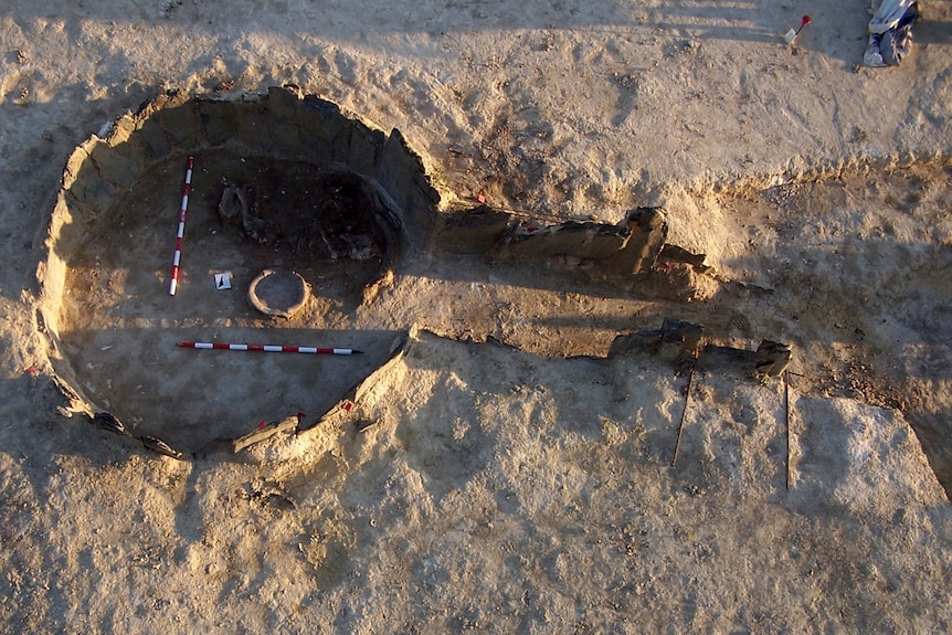 An aerial view of an open pre-historic circular tomb with an entrance corridor, showing measuring sticks.