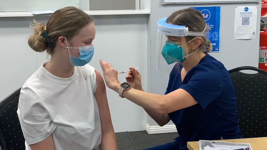 A nurse wearing a face shield provides a vaccination to the arm of a teenage girl.