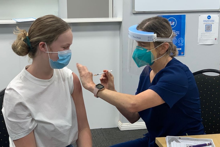 A nurse wearing a face shield provides a vaccination to the arm of a teenage girl.