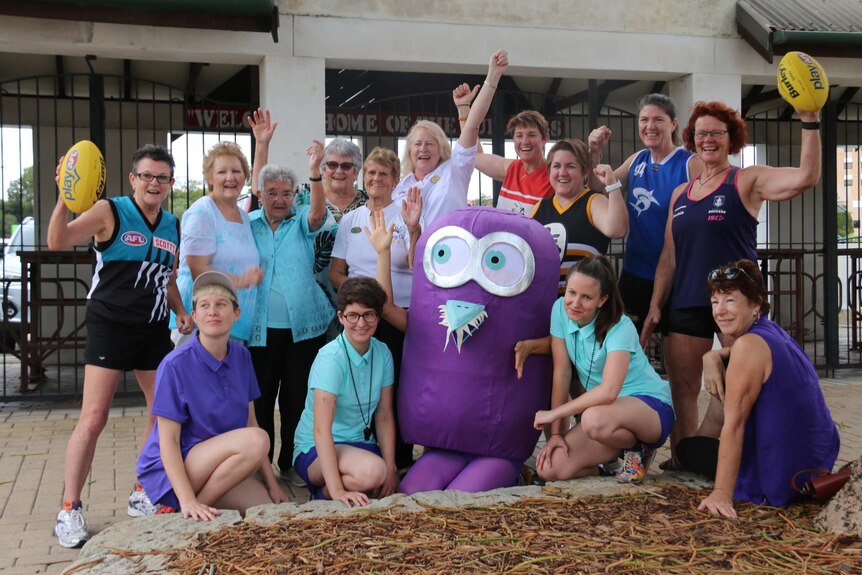 A group of about 15 women of various ages, some in football jumpers, pose for a photo with a purple mascot at Fremantle Oval.