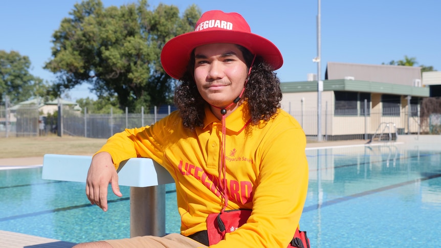 A man sits on the edge of a swimming pool wearing a yellow lifeguard shirt and red hat.