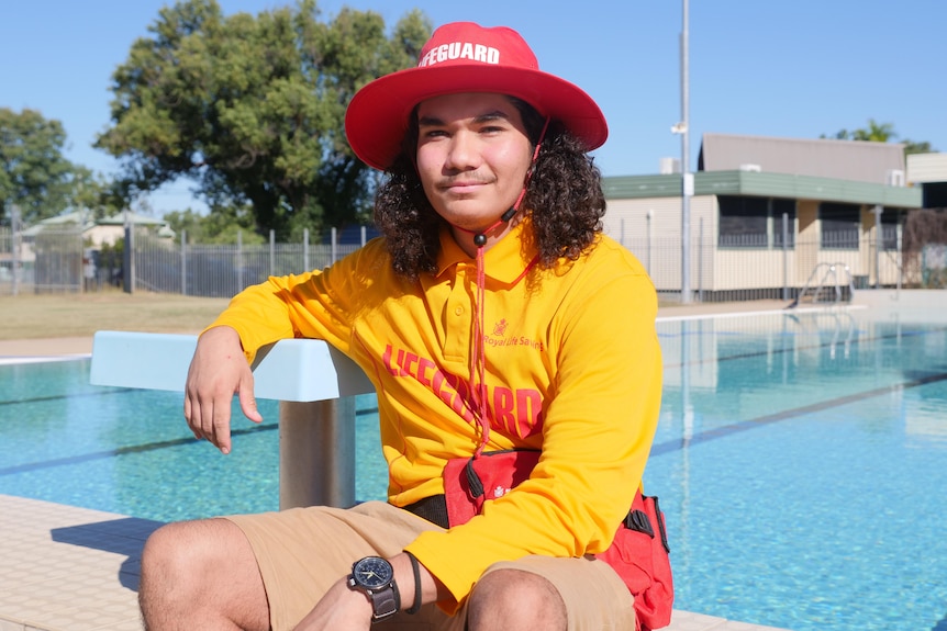 A man sits on the edge of a swimming pool wearing a yellow lifeguard shirt and red hat.