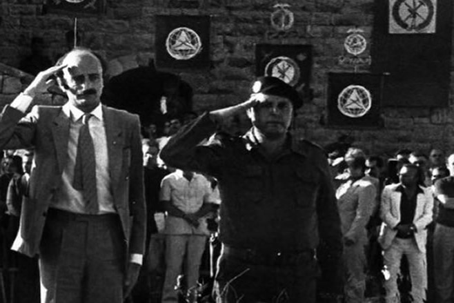 Walid Jumblatt in a black and white photo salutes, behind him are banners of his political party.