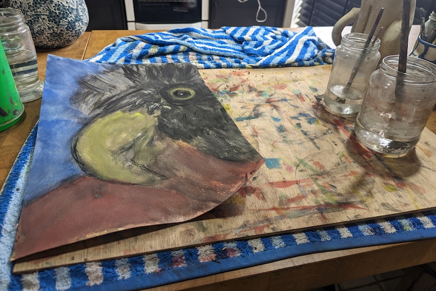 A watercolour painting of  bird is being prepared next to two glasses with brushes on a painting board.