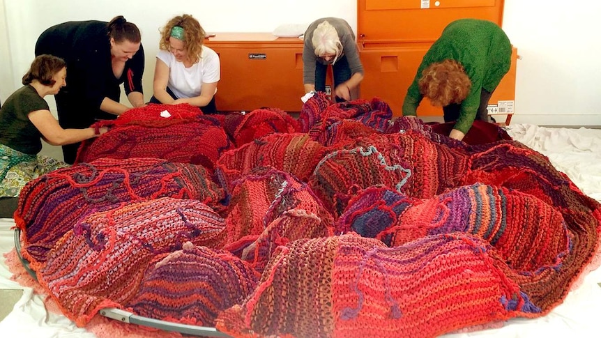 A group of women huddle together to knit a giant red placenta made of dyed t-shirts