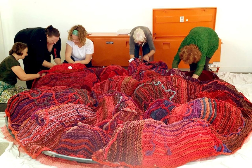 A group of women huddle together to knit a giant red placenta made of dyed t-shirts