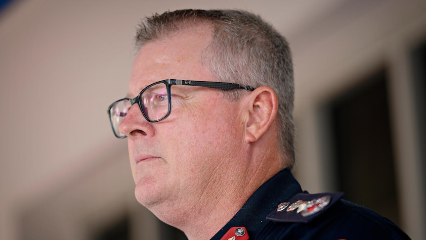 A man in a navy police uniform looks seriously off camera. He has grey hair and black glasses.