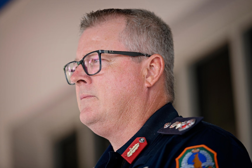 A man in a navy police uniform looks seriously off camera. He has grey hair and black glasses.