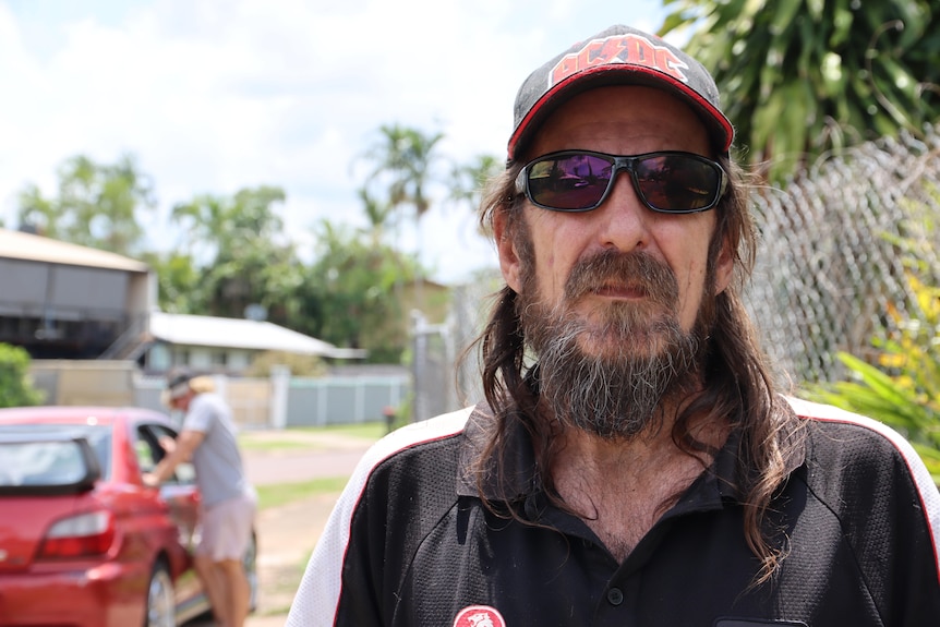 A man with long hair and in a ACDC cap, sunglasses and Holden T-shirt, standing on the side of a suburban street.