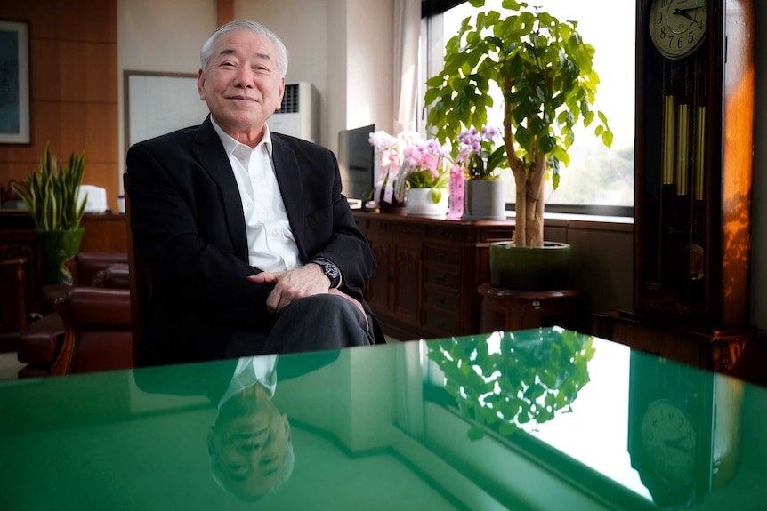 A Korean man in a suit sits cross legged and smiling behind a green desk