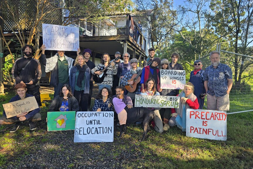 Twenty people with placards stand outside barricaded, flood-affected house.