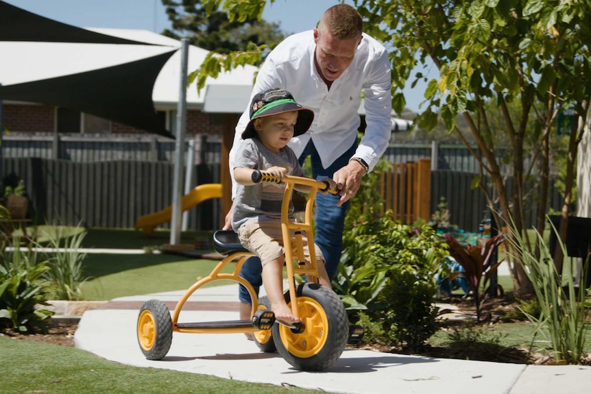 A man helps a kid ride a yellow tricycle down a path, which is surround by grass and plants.