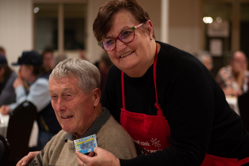 a bubbly woman with a red apron leans in over an elderly man, both are smiling.