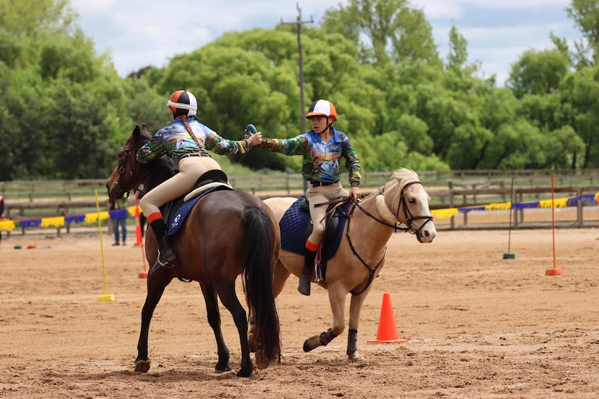 Two children riding brown ponies passed eachother. One of the children is handing a blue ring to the other.to each other.