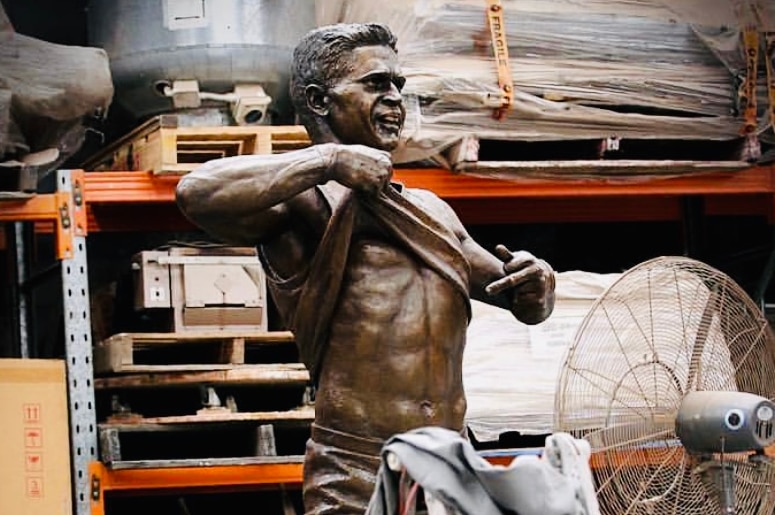 A bronze statue of a football player pulling up his top and pointing to his belly sits in a workshop.