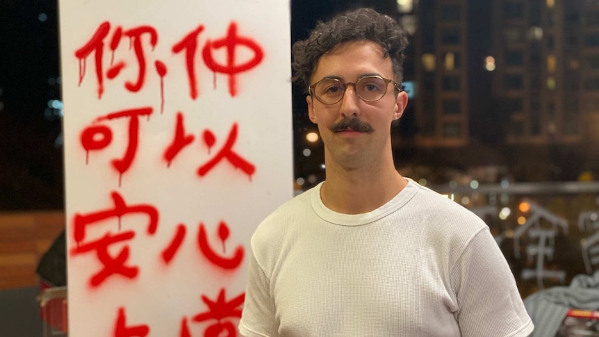 A man with a moustache and glasses stands in front of a pole with graffiti on it at night time.