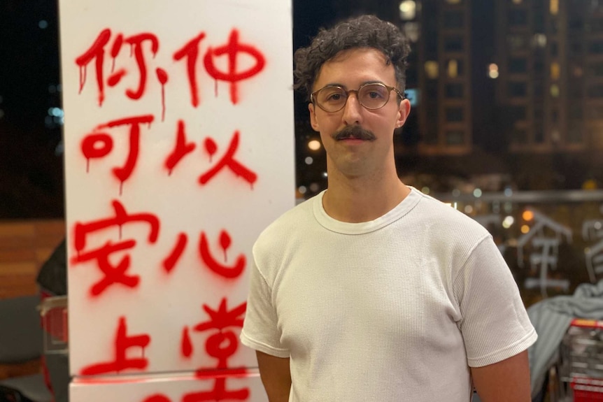 A man with a moustache and glasses stands in front of a pole with graffiti on it at night time.