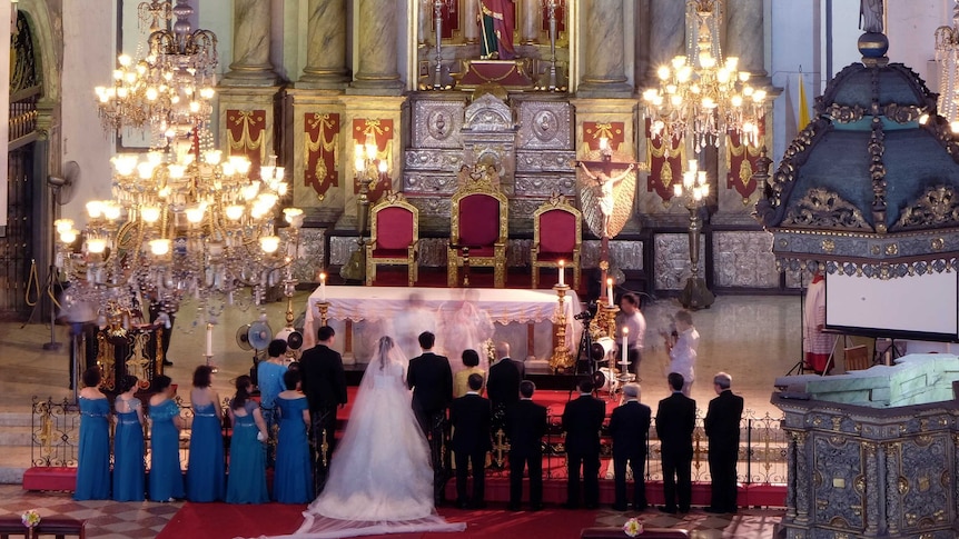 A man and a woman get married in a grand Christian church surrounded by bridesmaids and groomsmen.